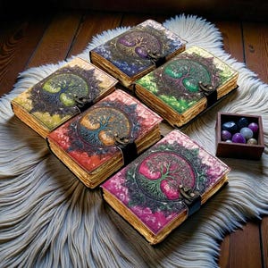 Puede incluir: Una colecci&oacute;n de cinco diarios encuadernados en cuero con cubiertas coloridas que presentan un dise&ntilde;o de &aacute;rbol de la vida. Cada diario tiene un cierre de metal y p&aacute;ginas envejecidas. Los diarios est&aacute;n colocados sobre una alfombra blanca y esponjosa.