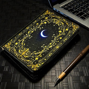 May include: A black book with a gold floral design and a glowing crescent moon. The book is next to a brown pen and a laptop. The cover has a detailed gold border and a starry background.
