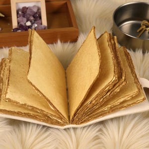 Puede incluir: Un diario hecho a mano abierto, con p&aacute;ginas de papel envejecido y texturizado. Las p&aacute;ginas son de un color marr&oacute;n dorado c&aacute;lido, y los bordes tienen un aspecto r&uacute;stico y desigual. El diario est&aacute; encuadernado en una cubierta blanca con una peque&ntilde;a pesta&ntilde;a decorativa.