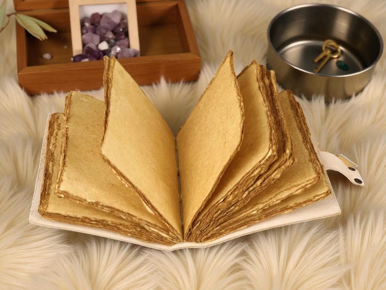 Puede incluir: Un diario hecho a mano abierto, con p&aacute;ginas de papel envejecido y texturizado. Las p&aacute;ginas son de un color marr&oacute;n dorado c&aacute;lido, y los bordes tienen un aspecto r&uacute;stico y desigual. El diario est&aacute; encuadernado en una cubierta blanca con una peque&ntilde;a pesta&ntilde;a decorativa.