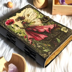 K&ouml;nnte beinhalten: Ein ledergebundenes Tagebuch mit einem Fantasy-Motiv auf dem Einband, das eine Fee in einem roten Kleid und mit gro&szlig;en Fl&uuml;geln zeigt, die eine rote Blume h&auml;lt. Das Tagebuch hat einen Metallverschluss und gen&auml;hte Kanten, mit gealterten Seiten.