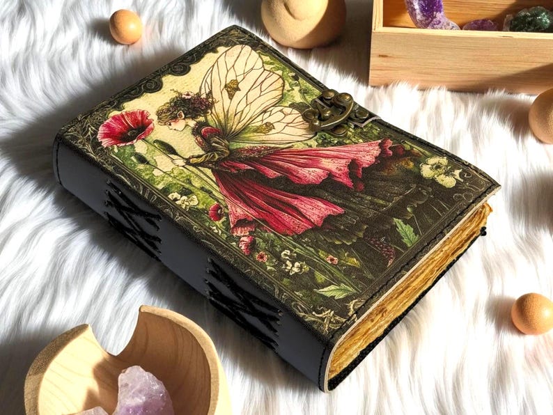 K&ouml;nnte beinhalten: Ein ledergebundenes Tagebuch mit einem Fantasy-Motiv auf dem Einband, das eine Fee in einem roten Kleid und mit gro&szlig;en Fl&uuml;geln zeigt, die eine rote Blume h&auml;lt. Das Tagebuch hat einen Metallverschluss und gen&auml;hte Kanten, mit gealterten Seiten.