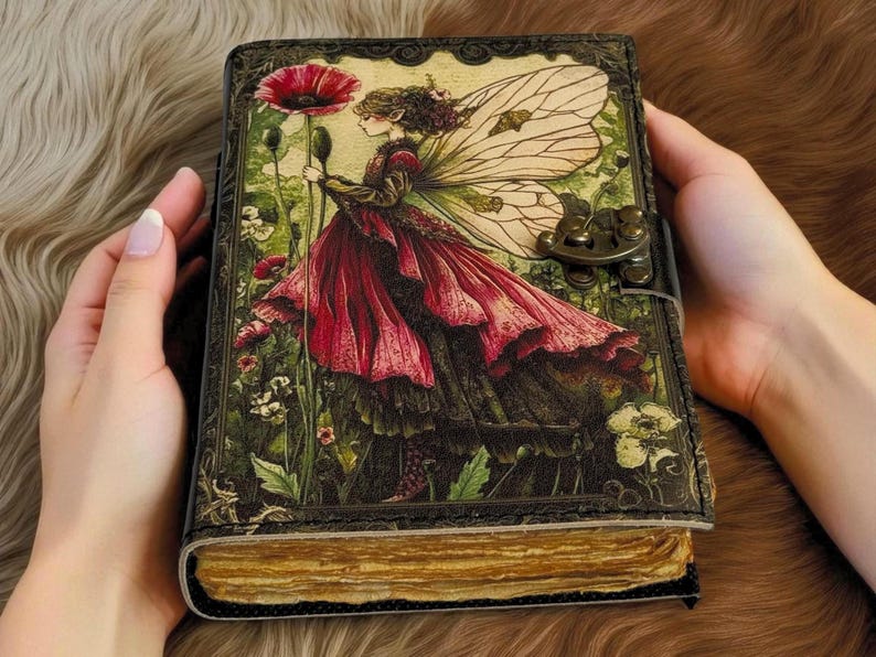 K&ouml;nnte beinhalten: Ein ledergebundenes Notizbuch mit einer Fantasy-Illustration auf dem Einband. Der Einband zeigt eine Fee in einem roten Kleid, die eine Mohnblume h&auml;lt. Das Notizbuch hat einen Metallverschluss und gealterte Seiten. Das Design hat eine Vintage- und skurrile &Auml;sthetik.