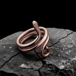 以下が含まれることがあります： 指に巻き付いた蛇の形をした銅色のリング。 蛇の頭は持ち上げられ、体は指の周りを螺旋状に巻いています。 リングは暗く、質感のある背景に配置されています。