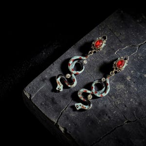 Puede incluir: Pendientes con forma de serpiente, con diseño esmaltado en azul claro y detalles rojos. Cada pendiente tiene una gema roja en la parte superior y pequeños cristales transparentes. Se presentan sobre una superficie oscura y texturizada.