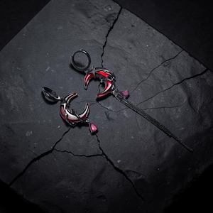 Puede incluir: Pendientes plateados con diseño de media luna. Las lunas tienen detalles de esmalte rojo y una gema rosa colgante. Un pendiente tiene una cadena larga y delgada con una gema rosa al final. Los pendientes se exhiben sobre una superficie oscura y texturizada.