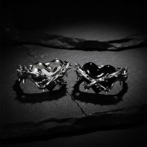 Puede incluir: Dos anillos con centros en forma de corazón. Uno tiene un corazón plateado, el otro un corazón negro. Ambos corazones están rodeados por una banda espinosa de color plateado. Los anillos se exhiben sobre una superficie oscura y texturizada.