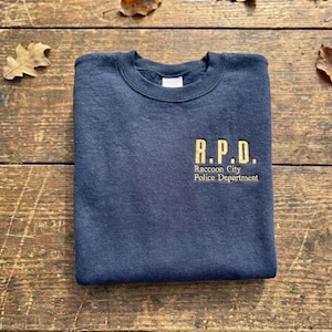 Könnte beinhalten: Ein marineblaues Sweatshirt mit goldfarbenem Text "R.P.D. Raccoon City Police Department". Das gefaltete Sweatshirt liegt auf einer Holzoberfläche, umgeben von Herbstblättern.