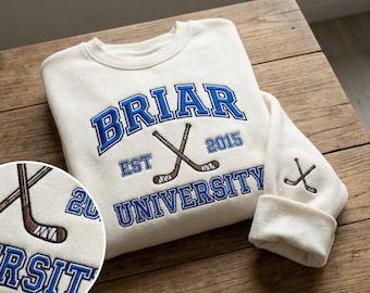 Op de afbeelding: Crèmekleurige sweatshirt met blauw geborduurde tekst: "BRIAR UNIVERSITY EST 2015." Voorzien van gekruiste hockeysticks. Een kleiner hockeystickontwerp staat op de mouwboord. De sweatshirt wordt gepresenteerd op een houten oppervlak.