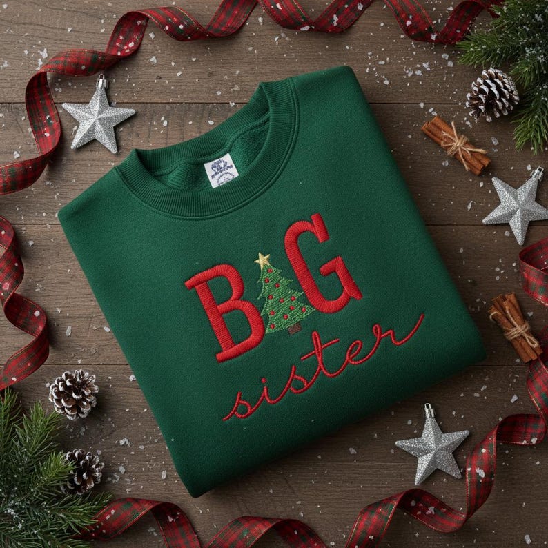 K&ouml;nnte beinhalten: Ein gr&uuml;nes Sweatshirt mit dem gestickten Text "BIG sister" in Rot. Das "G" zeigt ein Weihnachtsbaum-Design. Das Sweatshirt ist von Weihnachtsdekorationen umgeben, darunter B&auml;nder, Sterne, Tannenzapfen und Zimtstangen.