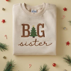 K&ouml;nnte beinhalten: Beiges Sweatshirt mit dem gestickten Schriftzug "BIG sister" in Braun. Das "I" in "BIG" ist durch ein Weihnachtsbaum-Design ersetzt. Das Bild ist mit Weihnachtsdekorationen geschm&uuml;ckt.