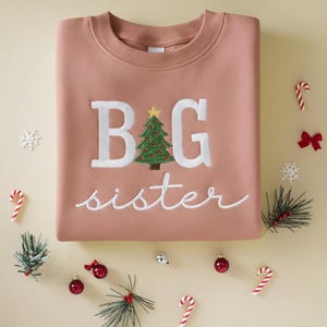 K&ouml;nnte beinhalten: Ein altrosa Sweatshirt mit wei&szlig; gesticktem Text "BIG sister" und einem Weihnachtsbaum-Motiv. Das Bild ist mit Zuckerstangen, Kugeln und Schneeflocken dekoriert.