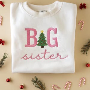 K&ouml;nnte beinhalten: Wei&szlig;es Sweatshirt mit rosa gesticktem Text &bdquo;B G sister&ldquo;. Zwischen dem &bdquo;B&ldquo; und dem &bdquo;G&ldquo; befindet sich ein gr&uuml;ner Weihnachtsbaum mit roten Ornamenten und einem gelben Stern. Das Sweatshirt ist von Weihnachtsdekorationen umgeben.