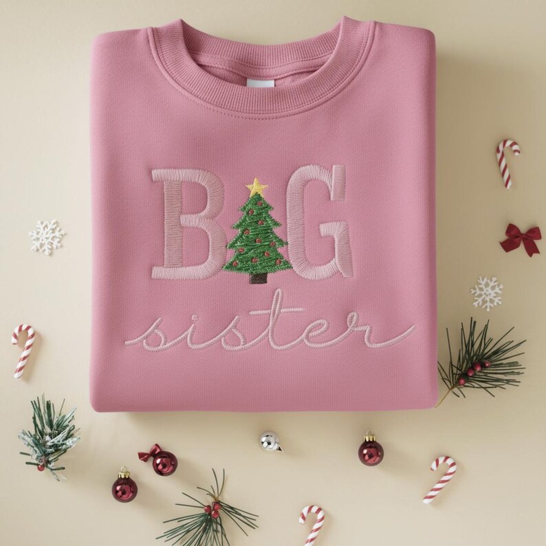 K&ouml;nnte beinhalten: Ein rosa Sweatshirt mit gesticktem Text und einem Weihnachtsbaum-Design. Der Text lautet "BIG sister" in einer Schreibschrift. Der Weihnachtsbaum ist gr&uuml;n mit roten Ornamenten und einem gelben Stern. Festliche Dekorationen umgeben das Sweatshirt.
