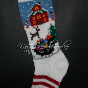 May include: A white knitted Christmas stocking with a blue and white striped cuff. The stocking features a red house with a green roof, a reindeer pulling a sleigh with presents, and a green Christmas tree. The stocking is decorated with white snowflakes and silver glitter.