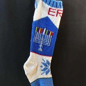 May include: A blue and white knitted stocking with a menorah and the word "ER" embroidered on it. The stocking has a white tassel at the top.
