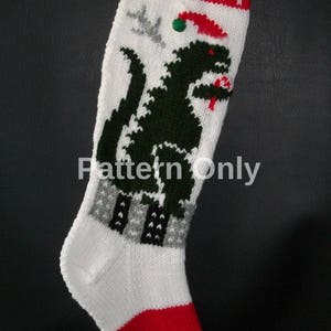 以下が含まれることがあります： 緑色の恐竜が赤いサンタの帽子をかぶった白いニットのクリスマスストッキング。ストッキングは赤いカフと緑色のつま先を持っています。ストッキングには「JONAT」という名前が編まれています。