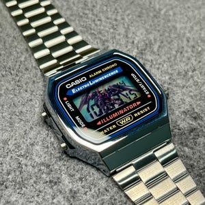 Puede incluir: Un reloj digital Casio plateado con una esfera rectangular y una correa de acero inoxidable. La esfera del reloj muestra la hora y presenta el texto "CASIO", "Electro Luminescence" e "ILLUMINATOR". El reloj es resistente al agua.