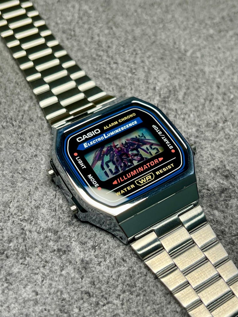 Puede incluir: Un reloj digital Casio plateado con una esfera rectangular y una correa de acero inoxidable. La esfera del reloj muestra la hora y presenta el texto "CASIO", "Electro Luminescence" e "ILLUMINATOR". El reloj es resistente al agua.