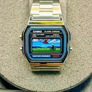 May include: A silver Casio digital watch with a black and blue face. The watch face displays a pixelated graphic of a plane and a motorcycle. The watch has a silver band and the text "CASIO ALARM CHRONO" is visible.