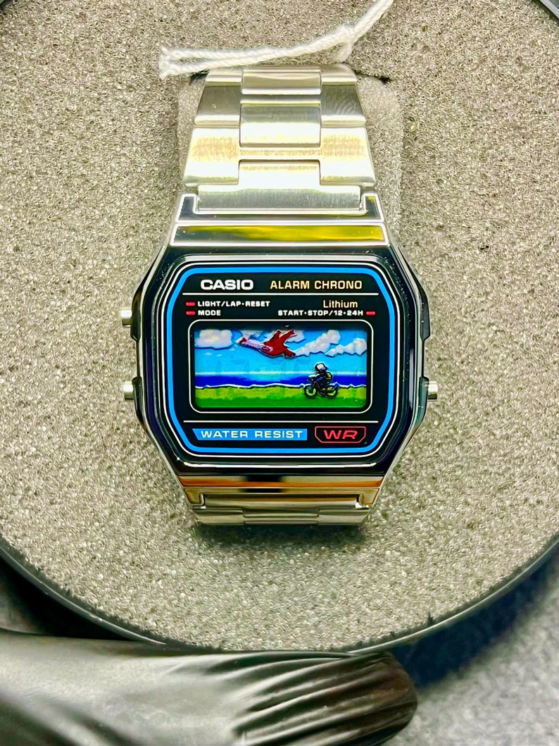 May include: A silver Casio digital watch with a black and blue face. The watch face displays a pixelated graphic of a plane and a motorcycle. The watch has a silver band and the text "CASIO ALARM CHRONO" is visible.