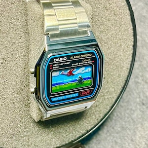 May include: A silver Casio digital watch with a black and blue display showing a pixelated graphic. The watch face has the text "CASIO ALARM CHRONO" and "WATER RESIST". The watch has a silver metal band and is displayed in a gray box.