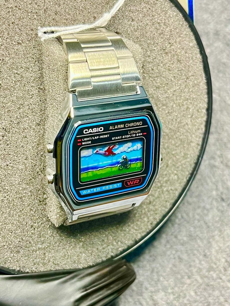 May include: A silver Casio digital watch with a black and blue display showing a pixelated graphic. The watch face has the text "CASIO ALARM CHRONO" and "WATER RESIST". The watch has a silver metal band and is displayed in a gray box.