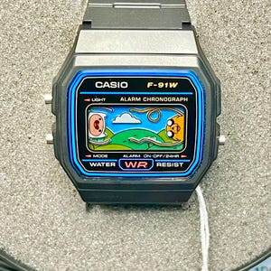 May include: Black Casio F-91W digital watch with a blue and black face featuring a cartoon scene. The watch has a black band and the face displays "CASIO F-91W", "LIGHT", "ALARM CHRONOGRAPH", "WATER RESIST".