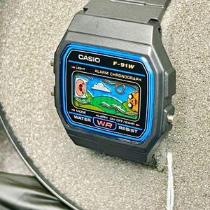 May include: A digital Casio F-91W watch with a black case and band. The watch face has a blue border and displays a cartoon graphic. The watch features light, alarm, and water resist functions. The watch is displayed on a textured surface.