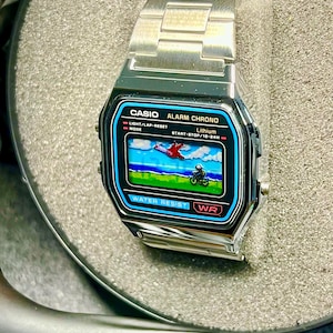 May include: A silver Casio digital watch with a rectangular face displaying a retro graphic of a plane and a motorcycle. The watch has a stainless steel band and features the text "CASIO ALARM CHRONO" and "WATER RESIST".