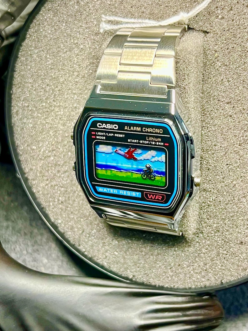 May include: A silver Casio digital watch with a rectangular face displaying a retro graphic of a plane and a motorcycle. The watch has a stainless steel band and features the text "CASIO ALARM CHRONO" and "WATER RESIST".