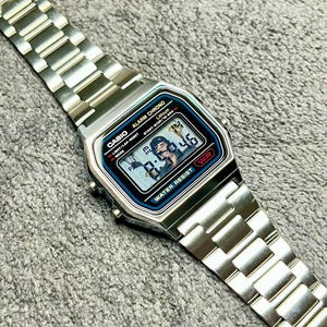 Può includere: Un orologio da polso digitale color argento con cinturino a maglie metalliche. Il quadrante rettangolare presenta un bordo nero con accenti blu, riportando il testo CASIO, ALARM CHRONO e WATER RESIST. Lo schermo digitale indica 8:46 PM.