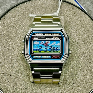 May include: A silver Casio digital watch with a rectangular face. The watch face displays a colorful graphic with the time 12:37:30. The watch has a stainless steel band and features the text "CASIO ALARM CHRONO" and "WATER RESIST WR".