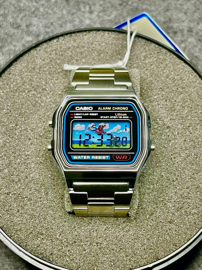 May include: A silver Casio digital watch with a rectangular face. The watch face displays a colorful graphic with the time 12:37:30. The watch has a stainless steel band and features the text "CASIO ALARM CHRONO" and "WATER RESIST WR".