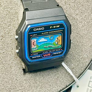 May include: A black Casio F-91W digital watch with a blue border around the screen. The watch face displays a cartoon scene. The watch is water resistant and has an alarm and chronograph functions. The watch is new in its original packaging.