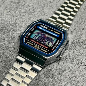 Puede incluir: Un reloj digital Casio plateado con esfera azul y negra. El reloj muestra la hora y tiene las palabras "CASIO" y "ELECTRO LUMINESCENCE" impresas en la esfera. La correa del reloj es de metal plateado.