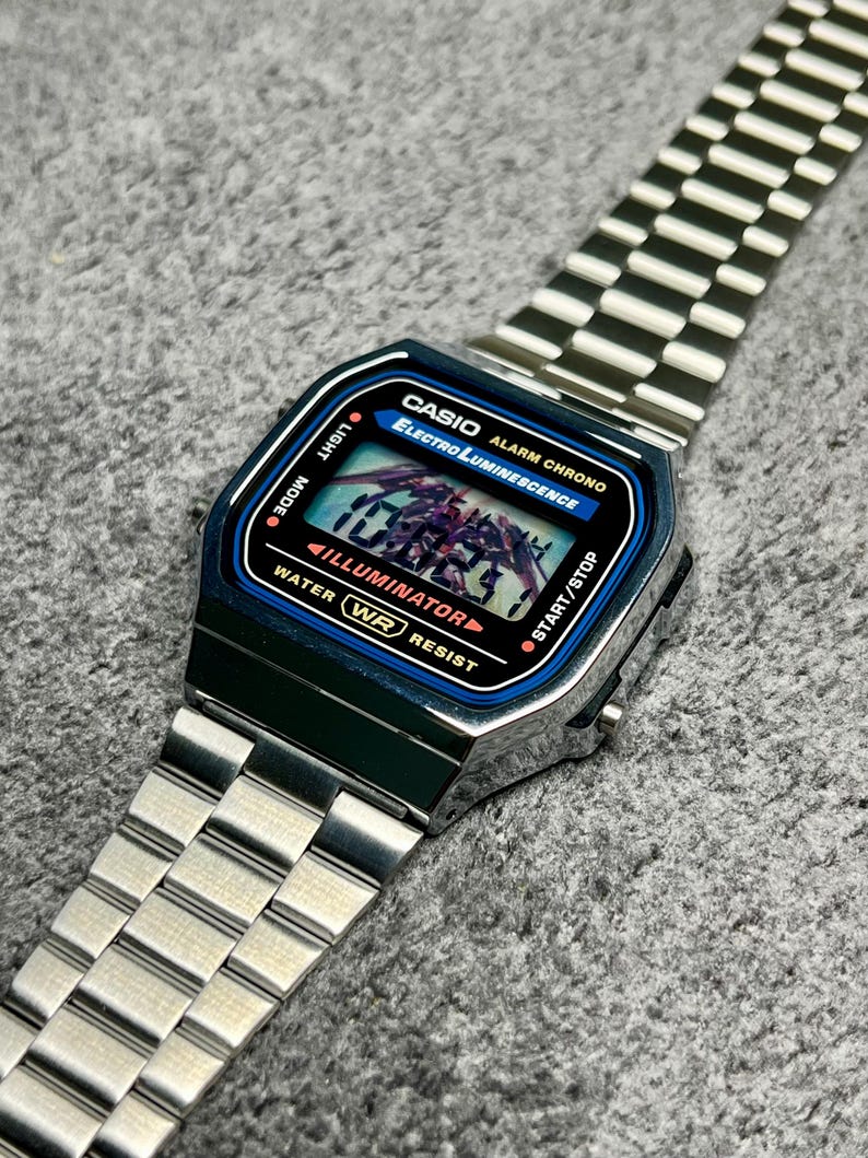 Puede incluir: Un reloj digital Casio plateado con esfera azul y negra. El reloj muestra la hora y tiene las palabras "CASIO" y "ELECTRO LUMINESCENCE" impresas en la esfera. La correa del reloj es de metal plateado.