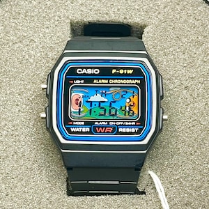 May include: A black Casio F-91W digital watch with a blue and white display. The watch face shows the time and has the text "CASIO F-91W". The watch is water resistant and has an alarm and light functions.