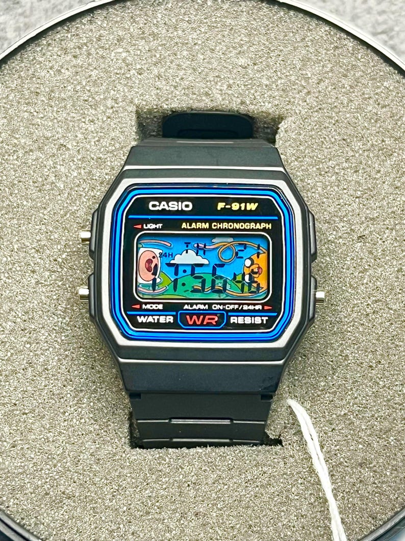May include: A black Casio F-91W digital watch with a blue and white display. The watch face shows the time and has the text "CASIO F-91W". The watch is water resistant and has an alarm and light functions.