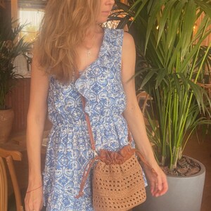 K&ouml;nnte beinhalten: Ein &auml;rmelloses Kleid mit blau-wei&szlig;em Muster, R&uuml;schenausschnitt und tailliertem Schnitt, kombiniert mit einer beigen H&auml;kel-Beuteltasche mit Kordelzug und einem braunen Schulterriemen in Lederoptik.