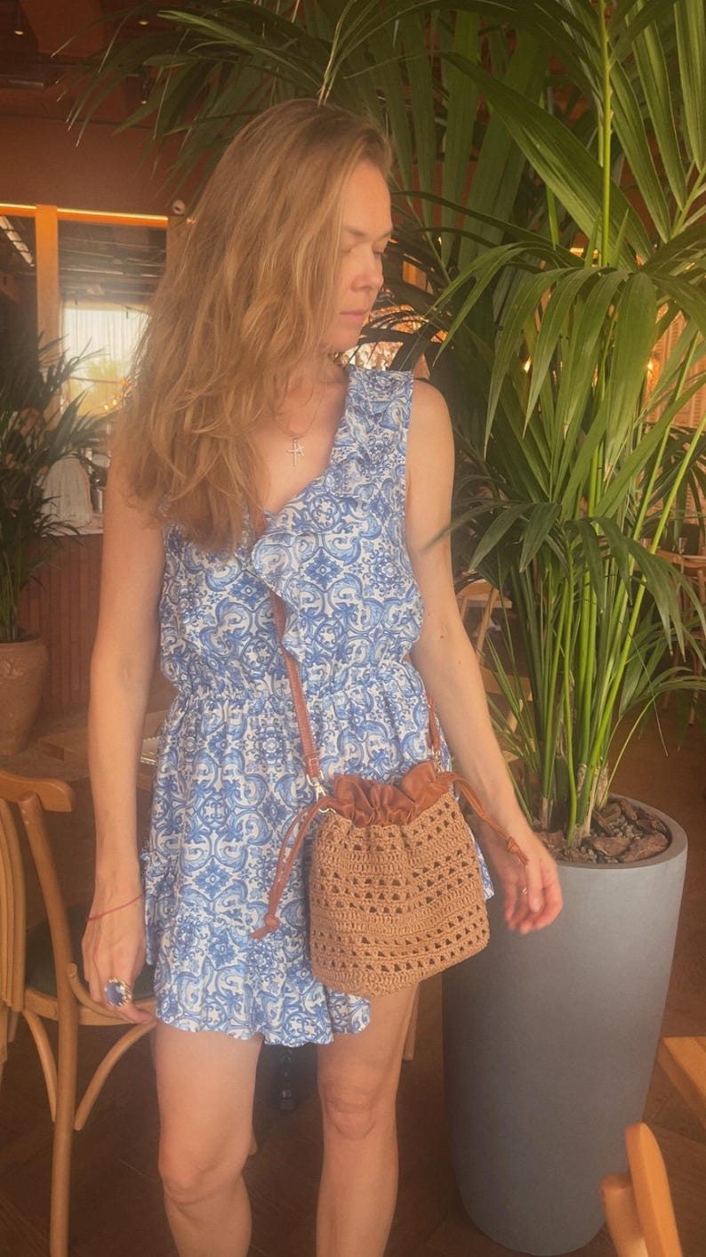 K&ouml;nnte beinhalten: Ein &auml;rmelloses Kleid mit blau-wei&szlig;em Muster, R&uuml;schenausschnitt und tailliertem Schnitt, kombiniert mit einer beigen H&auml;kel-Beuteltasche mit Kordelzug und einem braunen Schulterriemen in Lederoptik.