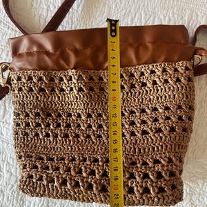 K&ouml;nnte beinhalten: Eine Beuteltasche aus braunem Kunstleder und geflochtenem Stroh mit Kordelzug und langem Schulterriemen. Ein gelbes Ma&szlig;band liegt vertikal am Beutel an und zeigt eine H&ouml;he von etwa 24 cm von der Unterseite bis zur oberen Kante.