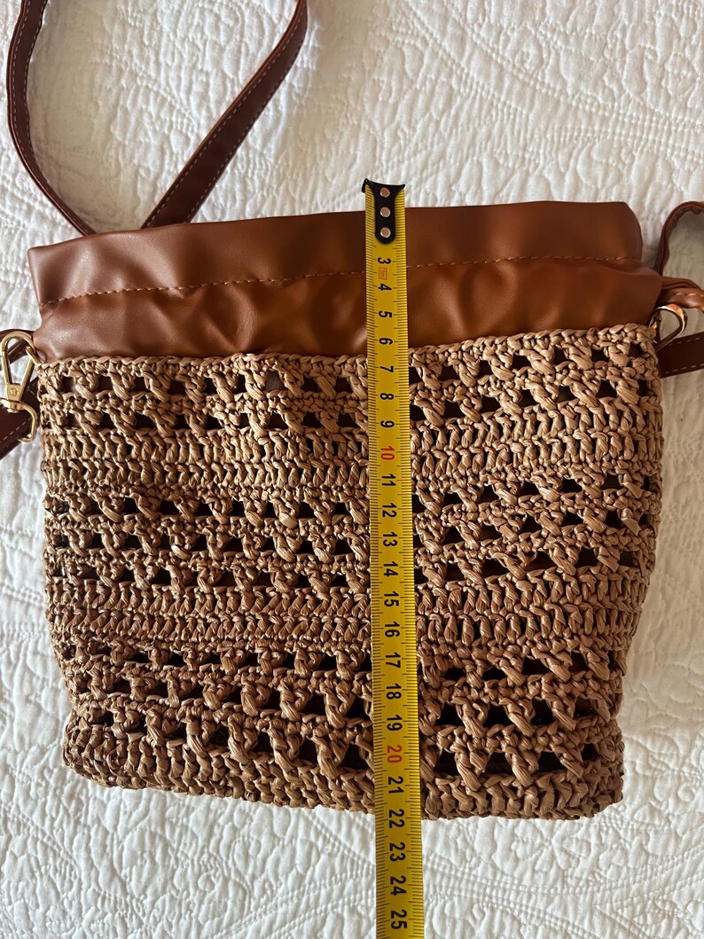 K&ouml;nnte beinhalten: Eine Beuteltasche aus braunem Kunstleder und geflochtenem Stroh mit Kordelzug und langem Schulterriemen. Ein gelbes Ma&szlig;band liegt vertikal am Beutel an und zeigt eine H&ouml;he von etwa 24 cm von der Unterseite bis zur oberen Kante.