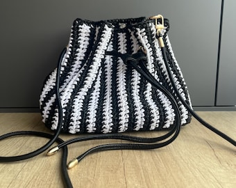 Könnte beinhalten: Eine Beuteltasche mit einem Häkelmuster aus abwechselnden vertikalen schwarzen und weißen Streifen. Die Tasche verfügt über einen schwarzen Kordelzug mit goldfarbenen Metallenden und einen langen, dünnen schwarzen Schulterriemen mit goldenen Beschlägen.