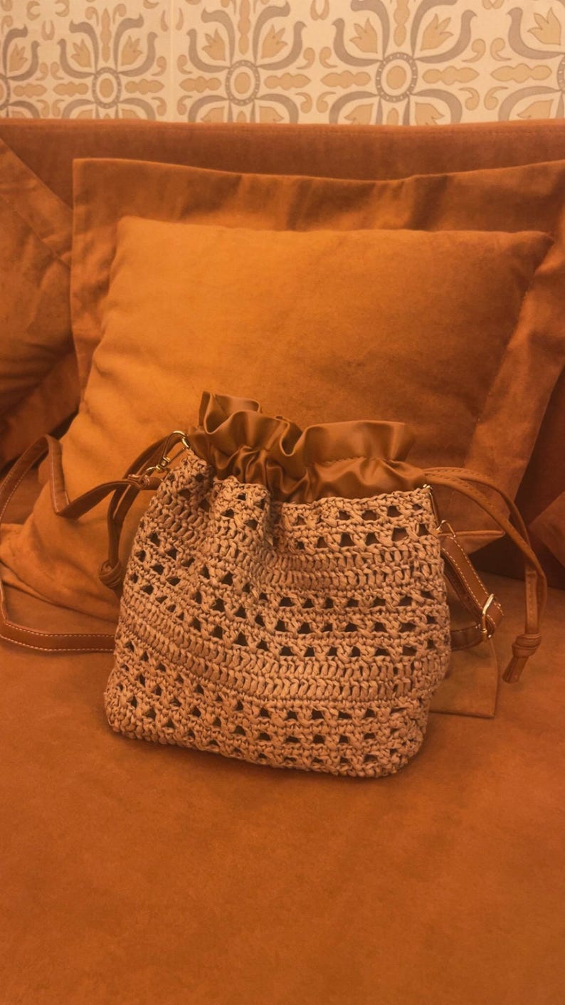 K&ouml;nnte beinhalten: Eine beige H&auml;kel-Beuteltasche mit einem durchbrochenen Muster und einem glatten, einfarbigen braunen Kordelzugverschluss an der Oberseite. Die Tasche verf&uuml;gt &uuml;ber einen passenden braunen Schulterriemen mit goldfarbenen Metallbeschl&auml;gen.