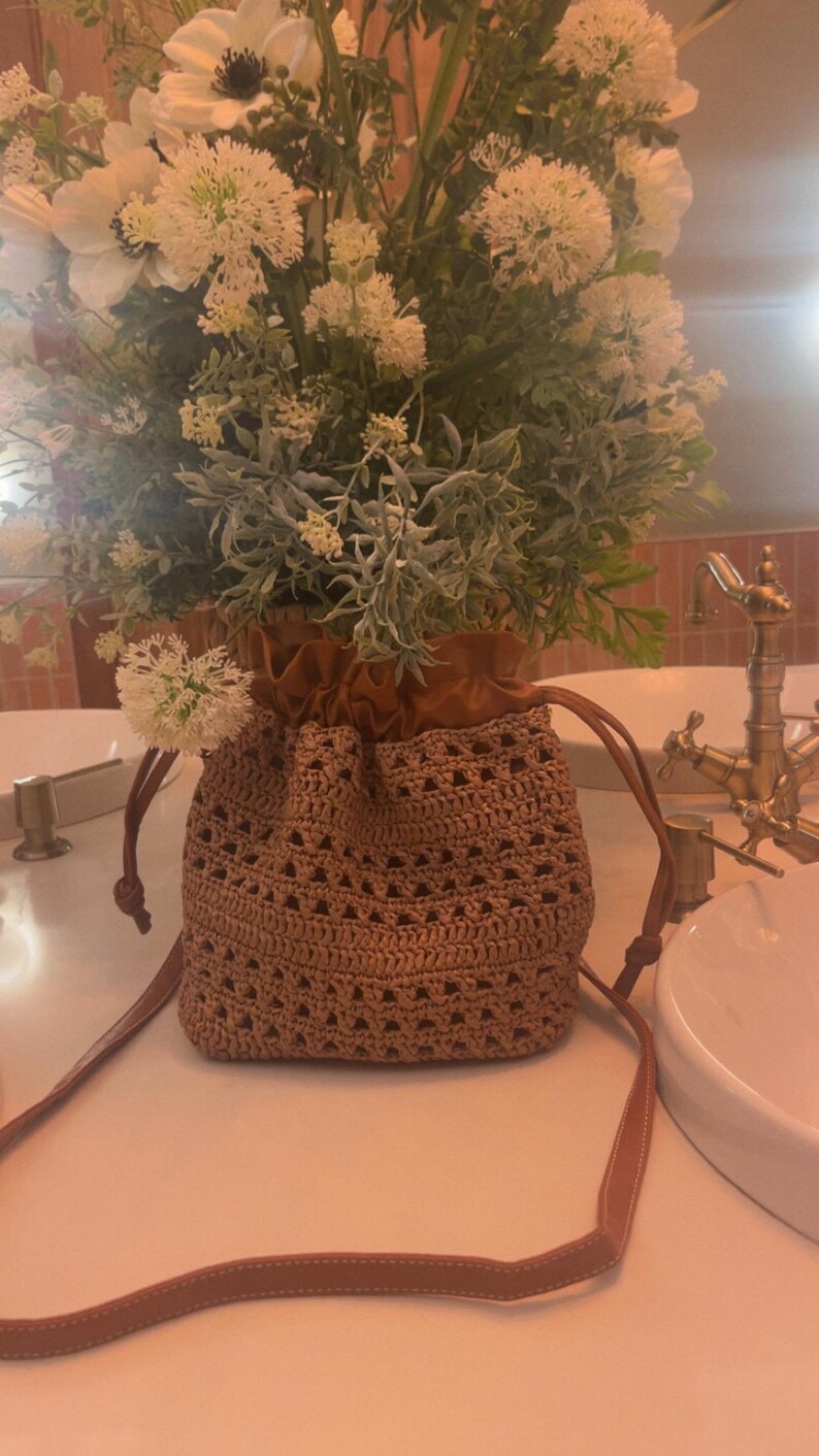 K&ouml;nnte beinhalten: Eine beigefarbene H&auml;kel-Beuteltasche mit dreieckigem Lochmuster und einem oberen Abschnitt aus glattem braunem Stoff. Die Tasche hat lange, d&uuml;nne braune Riemen und ist mit einem gro&szlig;en Arrangement aus wei&szlig;en Blumen und gr&uuml;nem Blattwerk gef&uuml;llt.