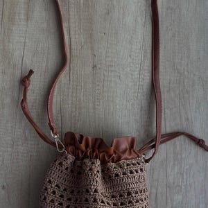K&ouml;nnte beinhalten: Eine Beuteltasche mit Kordelzug, bestehend aus einem braunen H&auml;kelk&ouml;rper mit offenem geometrischem Muster. Die Tasche verf&uuml;gt &uuml;ber einen glatten braunen Kunstleder-Oberteil mit gerafftem Verschluss und einen passenden d&uuml;nnen braunen Schulterriemen mit Metallbeschl&auml;gen.