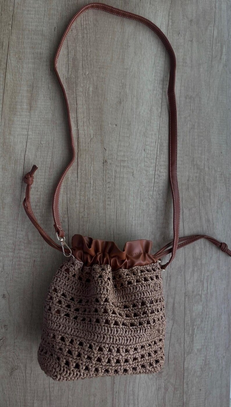 K&ouml;nnte beinhalten: Eine Beuteltasche mit Kordelzug, bestehend aus einem braunen H&auml;kelk&ouml;rper mit offenem geometrischem Muster. Die Tasche verf&uuml;gt &uuml;ber einen glatten braunen Kunstleder-Oberteil mit gerafftem Verschluss und einen passenden d&uuml;nnen braunen Schulterriemen mit Metallbeschl&auml;gen.