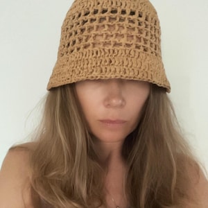 Pode incluir: Um chapéu bucket bege feito com técnica de crochê, apresentando um padrão de trama aberta ao redor da copa e uma aba sólida e densamente tecida. O chapéu é construído a partir de um material de fio fosco e texturizado em tom castanho.