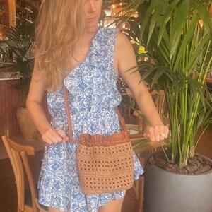K&ouml;nnte beinhalten: Eine beige gewebte Beuteltasche mit Kordelzugverschluss und einem braunen Lederriemen. Die Tasche zeichnet sich durch ein strukturiertes Strickmuster am gesamten K&ouml;rper und einen glatten Lederabschnitt oben mit einem Kordelzugmechanismus aus.